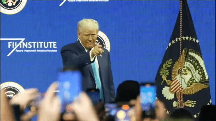 President Donald Trump delivering remarks at the FII Summit in Miami, standing on stage with the presidential seal and audience members capturing the moment on their smartphones.